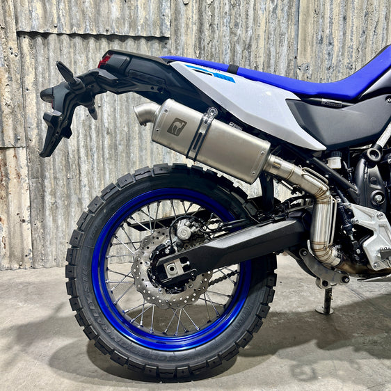 A Yamaha 700 Tenere motorcycle with a n Enduro bend high exhaust system by Camel ADV Products, displayed indoors with a logo in the background.